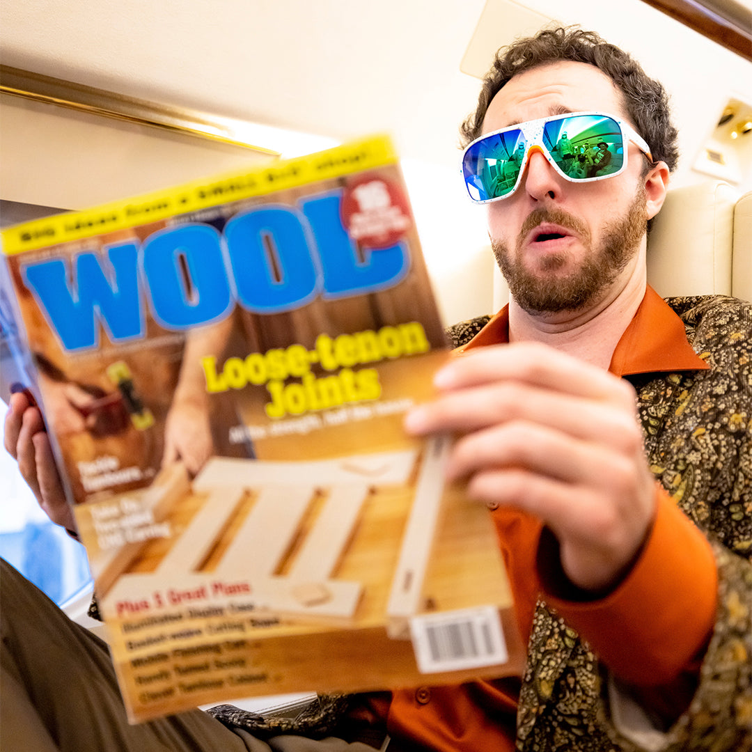 / Blue-Green | A man reading a magazine on a plane wearing Pit Viper Sunglasses